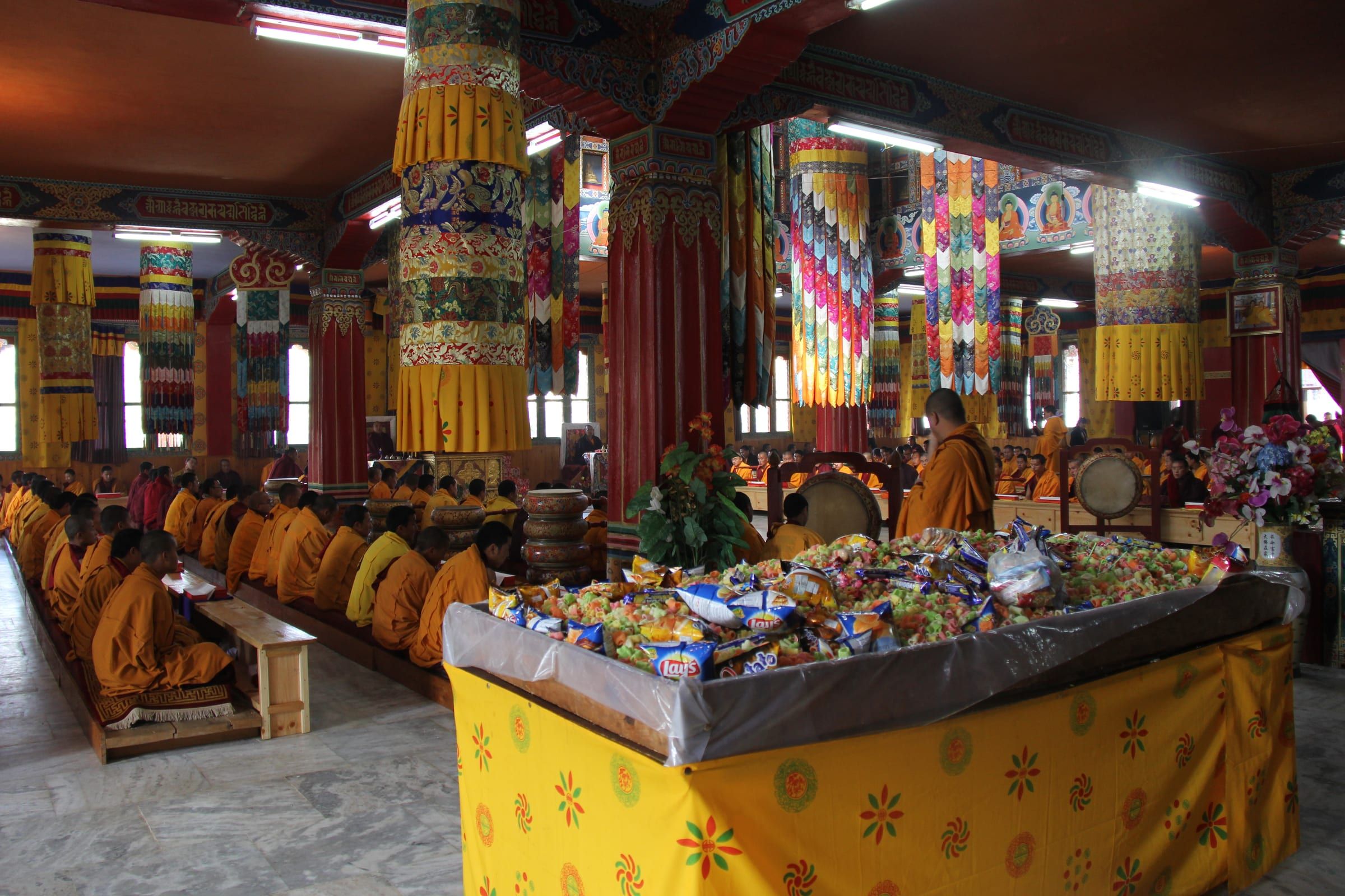 Monks in rows chant in a painted temple hall behind a table heaped with offerings.