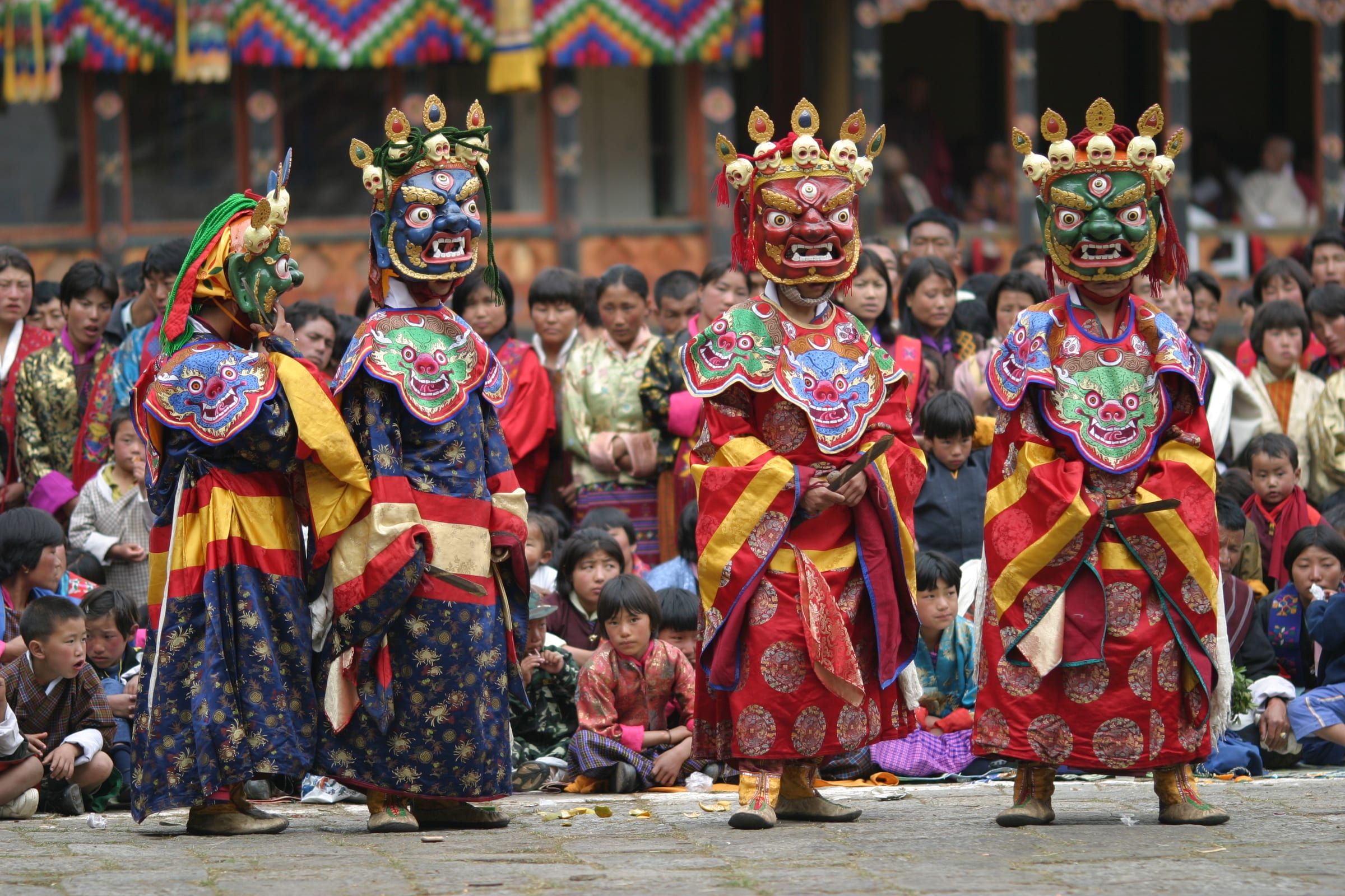 Masked dancers in red and blue brocade stand in a line before a seated crowd.