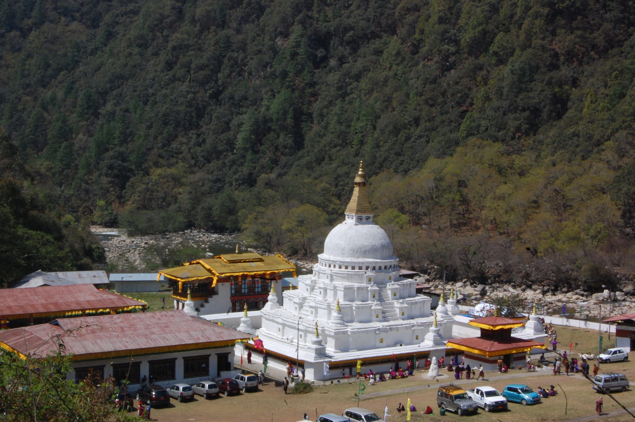 Chorten Kora, a large white stupa, beside a river below a forested hillside.