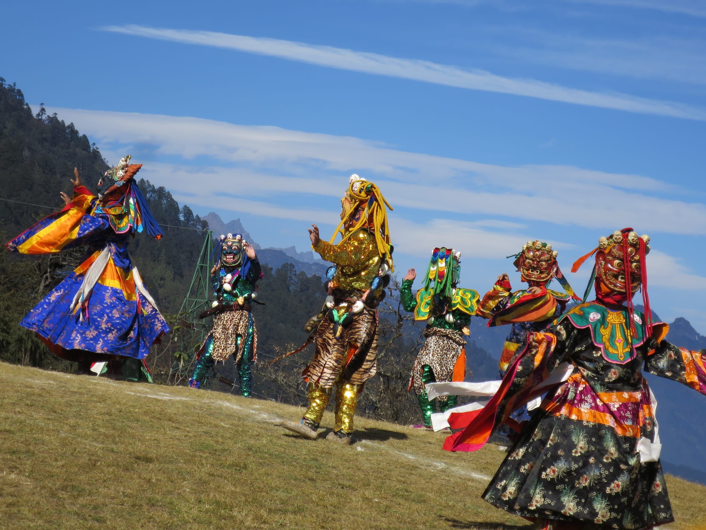 Masked dancers in brocade robes on an open grassy ridge under a blue sky.