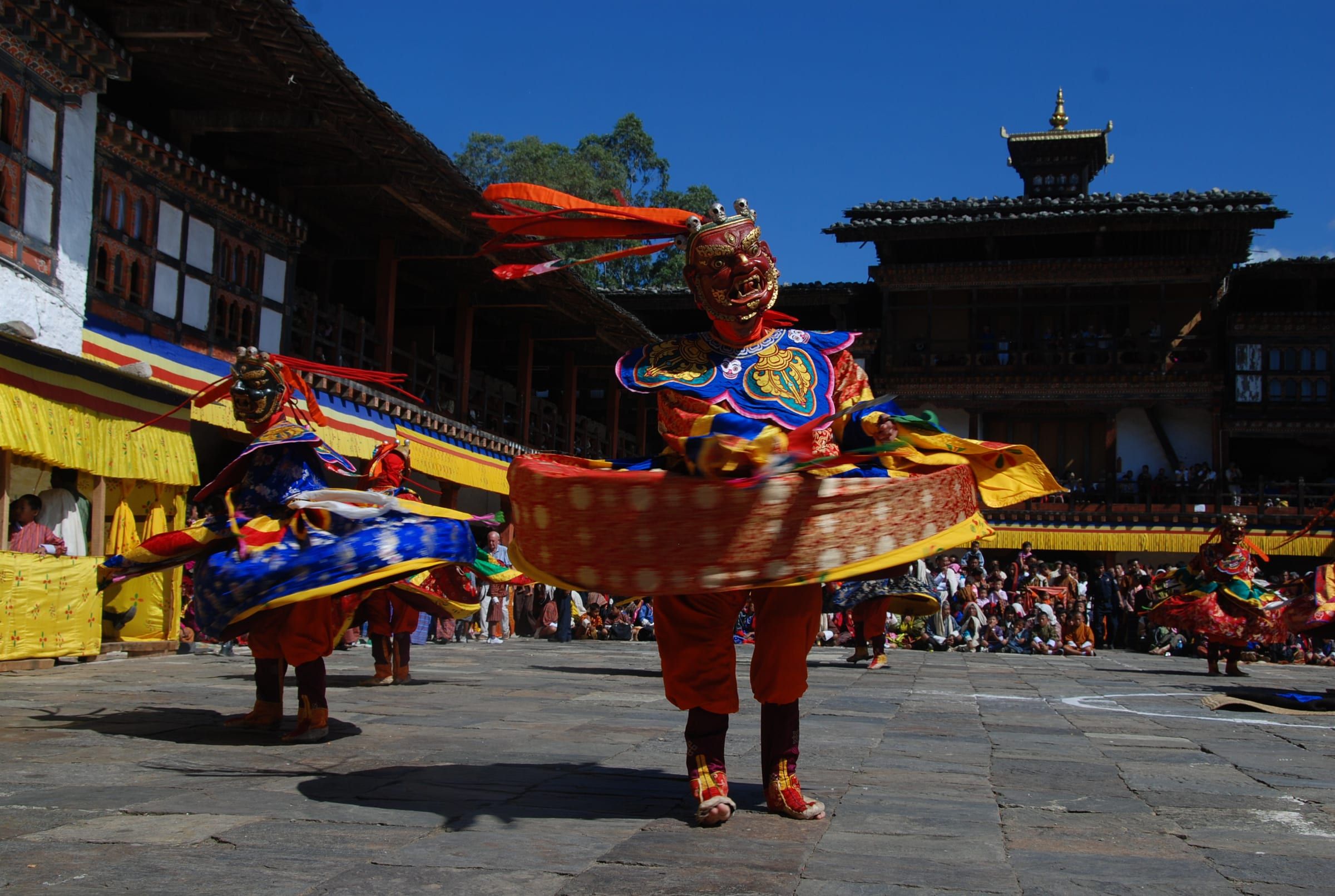 A masked dancer spins in a flagstone courtyard ringed by wooden galleries.