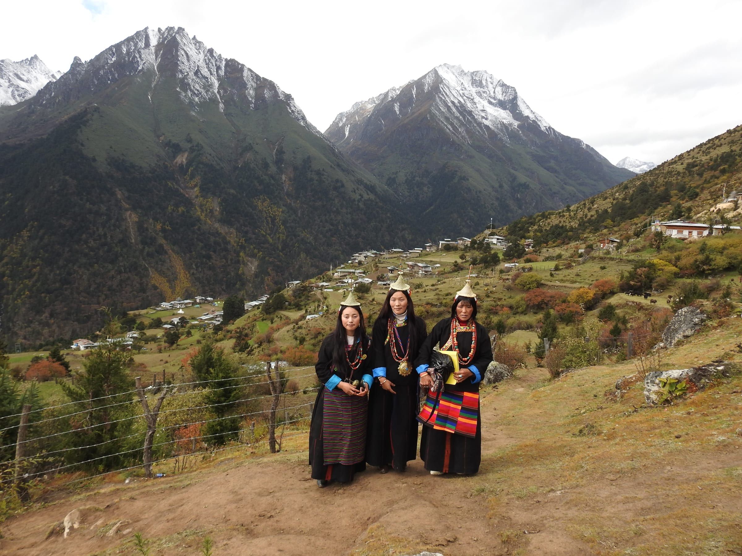 Three women in pointed bamboo hats on a hillside trail below snow-dusted peaks.