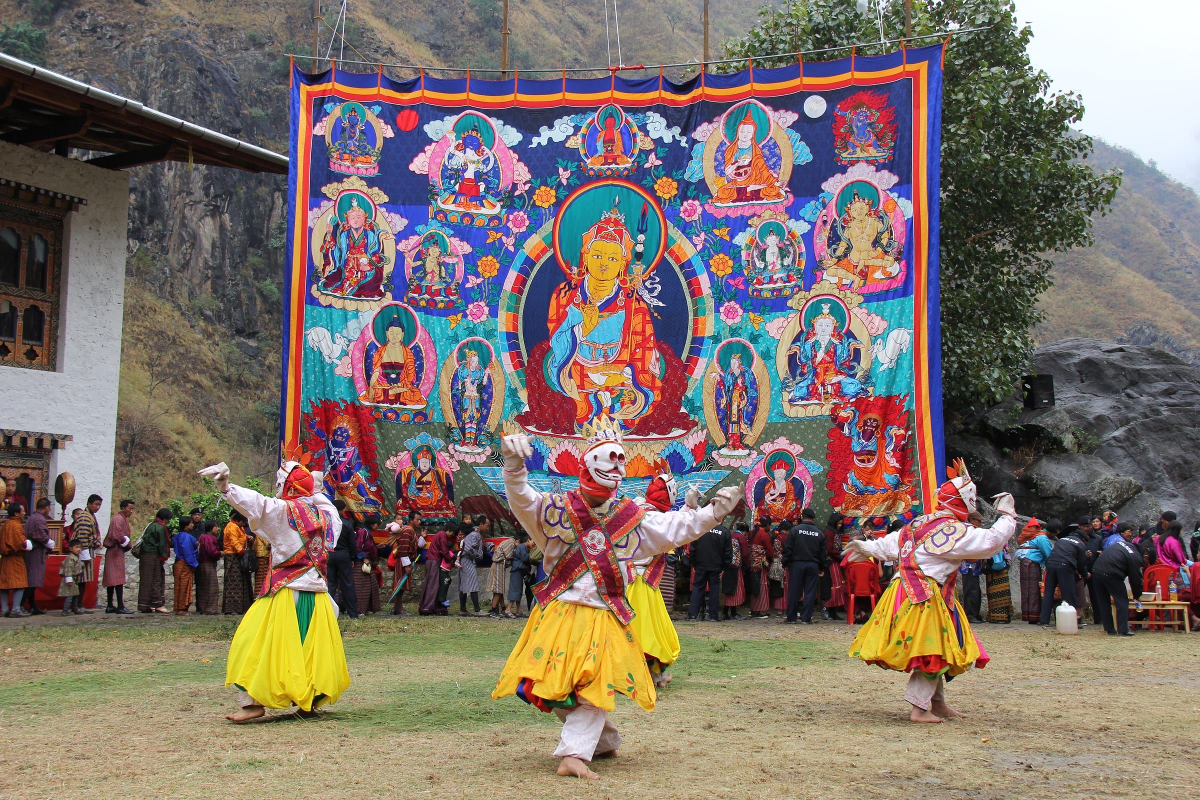 Masked dancers in yellow skirts in front of a giant thongdrel of Guru Rinpoche.
