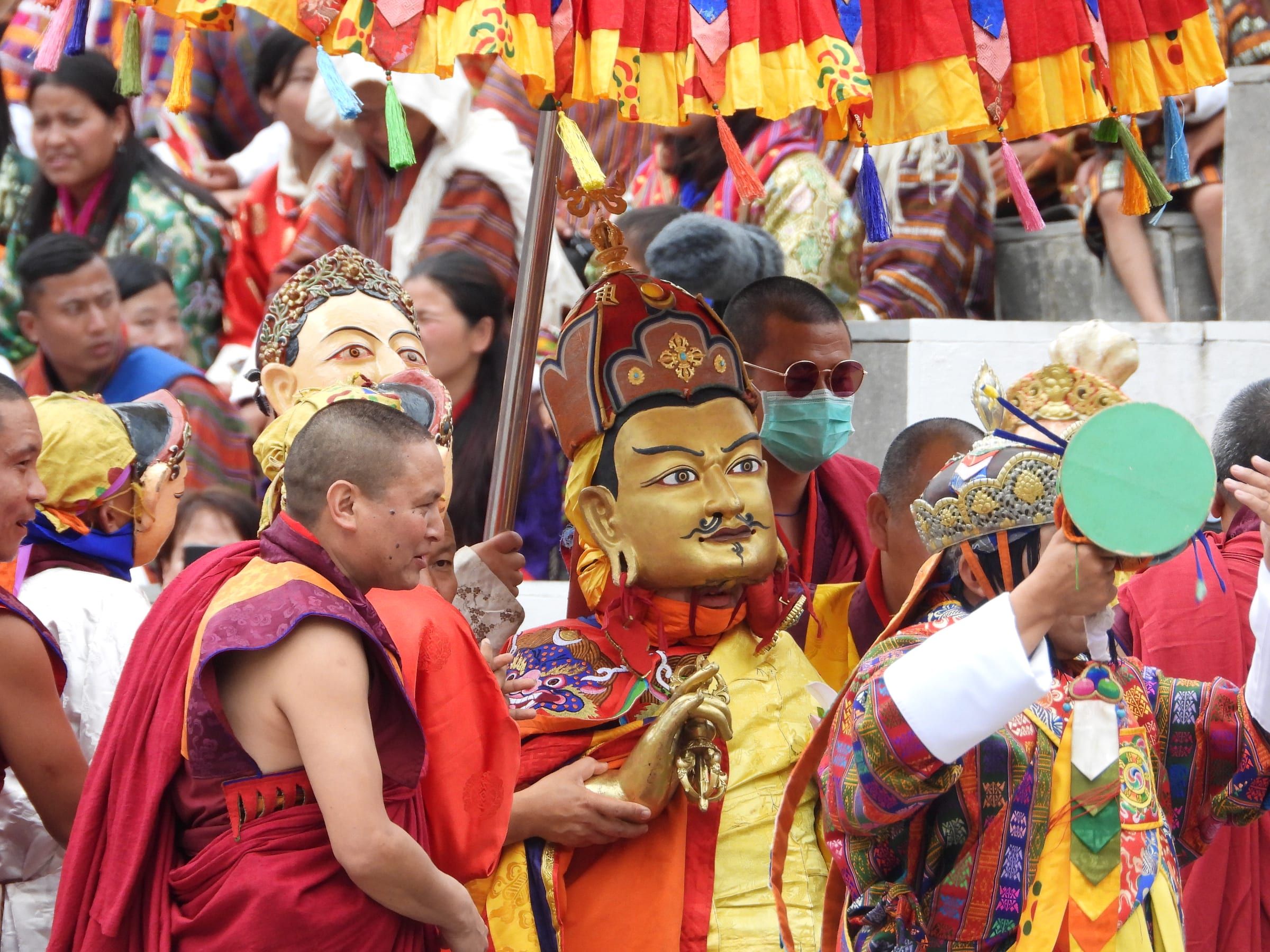 A dancer in the golden mask of Guru Rinpoche walks under a fringed parasol among monks and masked attendants.