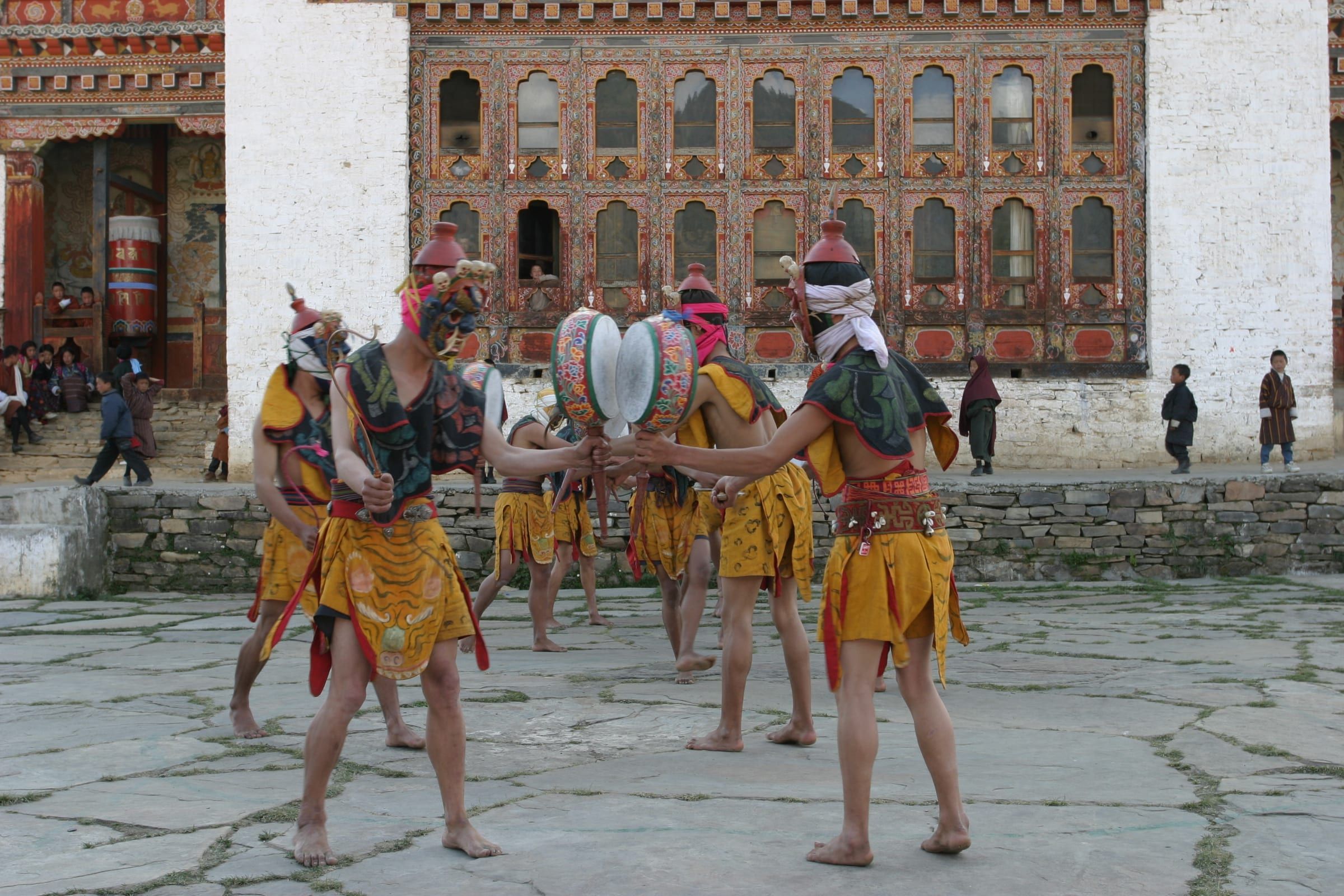 Masked dancers with hand drums in a stone courtyard in front of a temple.