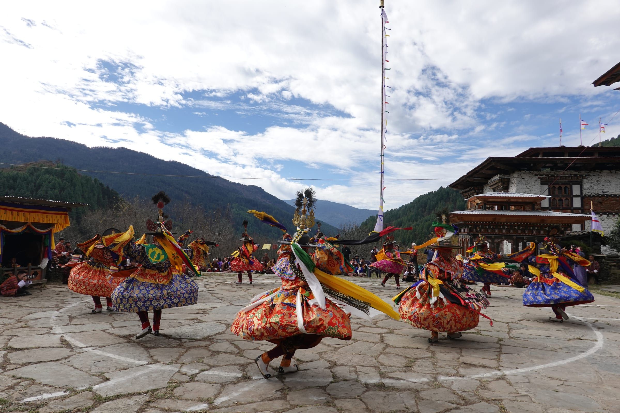 Masked dancers in silk costumes spin in a circle, with forested mountains behind.