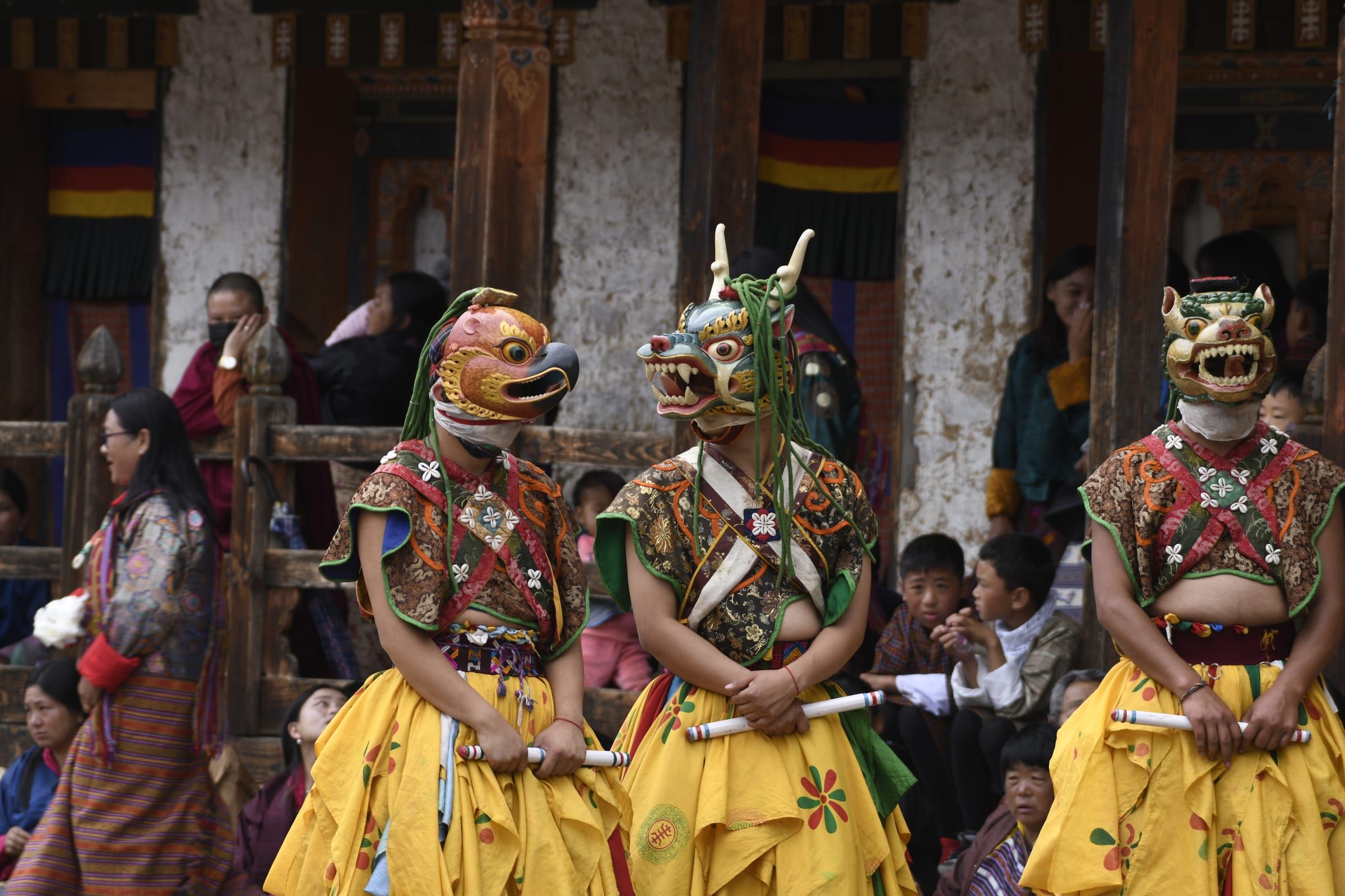 Three masked dancers in ritual dress stand among onlookers in a temple courtyard.