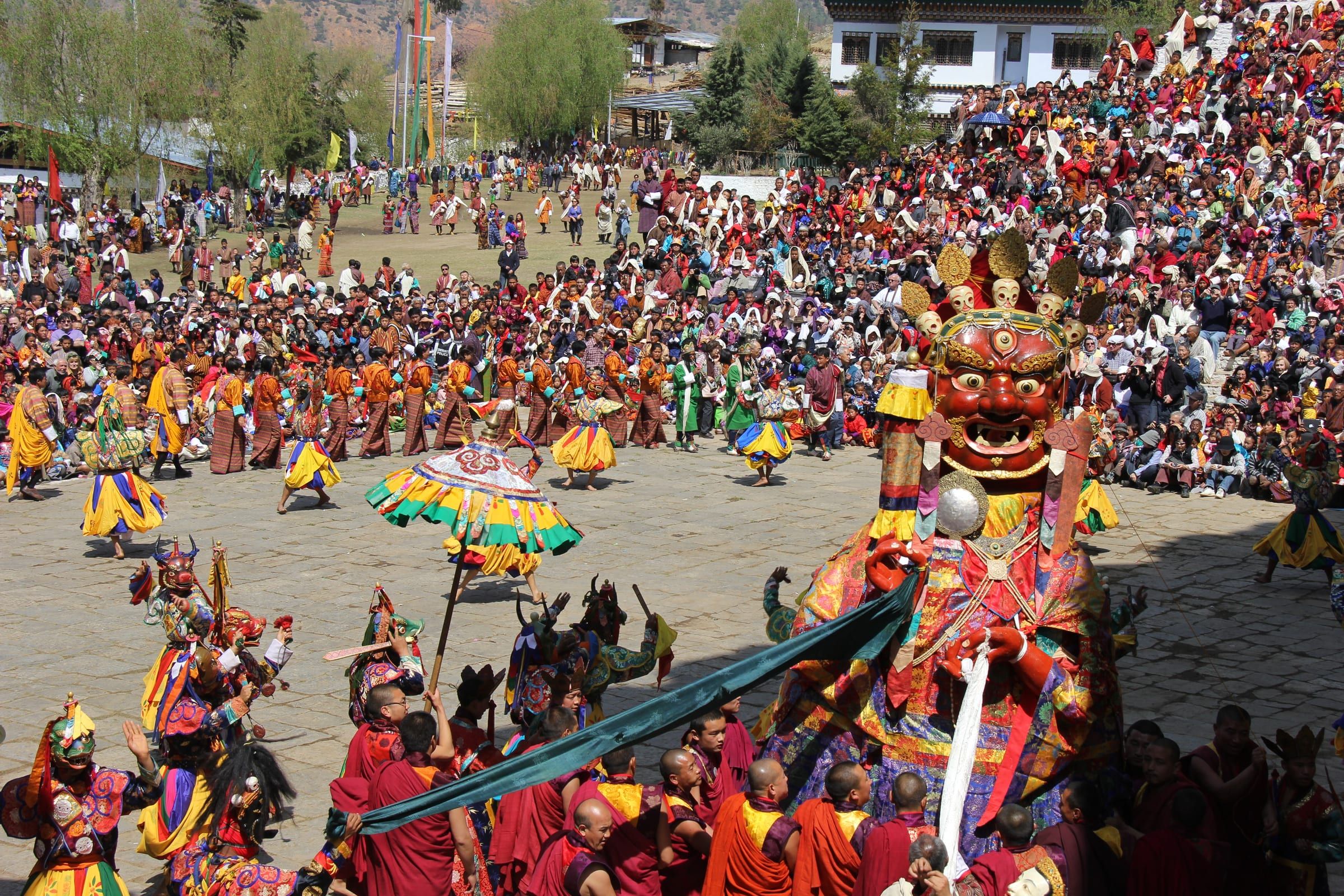 Monks guide a giant masked deity figure through a crowded courtyard, with masked dancers around it.
