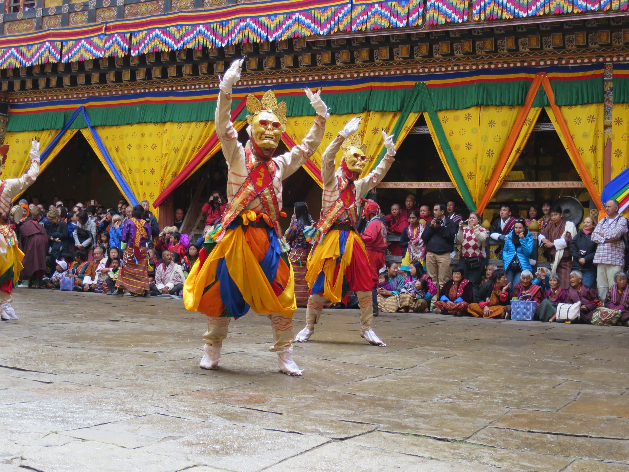 Masked dancers perform in a dzong courtyard before a crowd under coloured canopies.