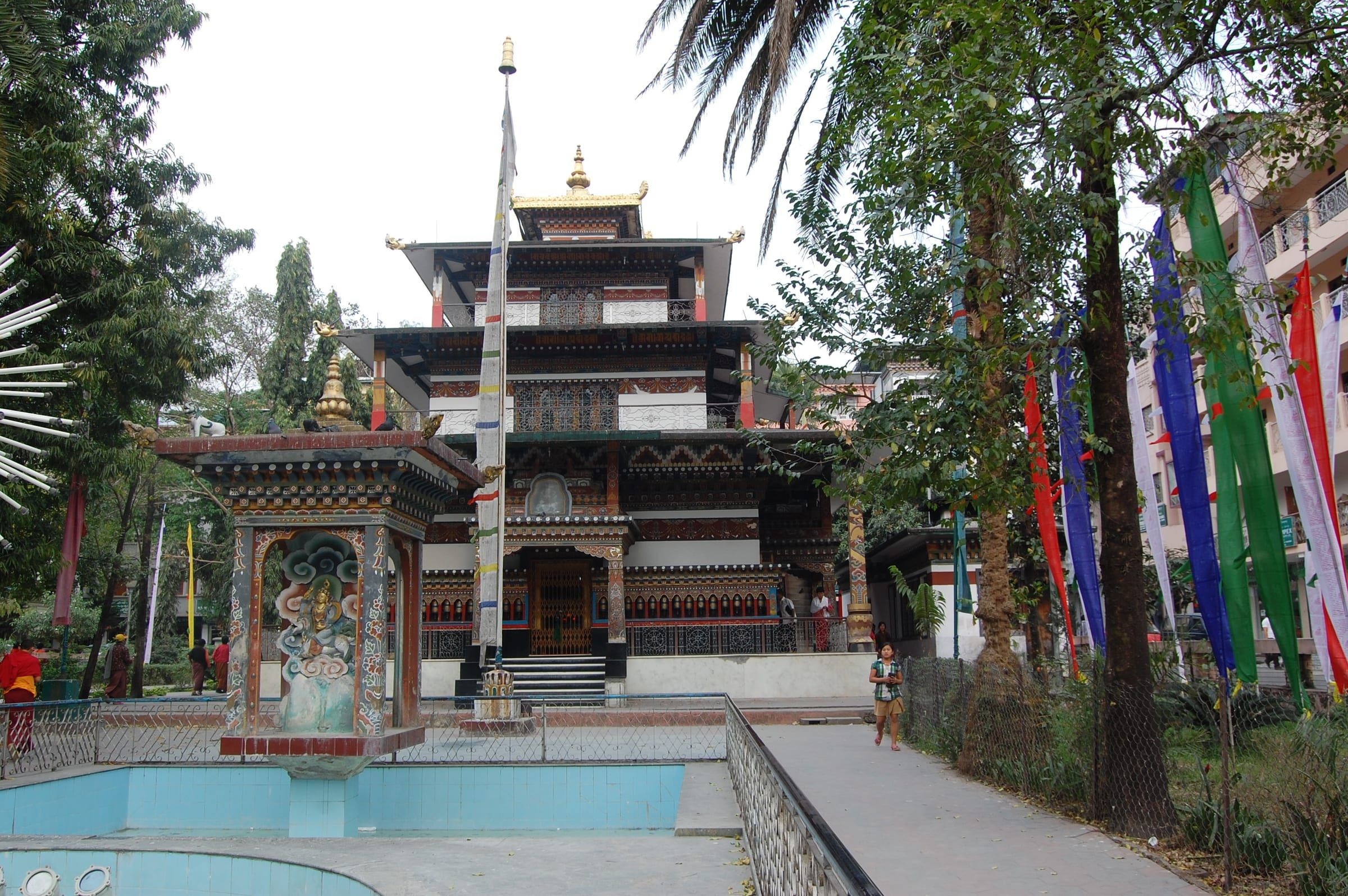 A tiered temple behind a small shrine and a path lined with prayer flags and palm trees.