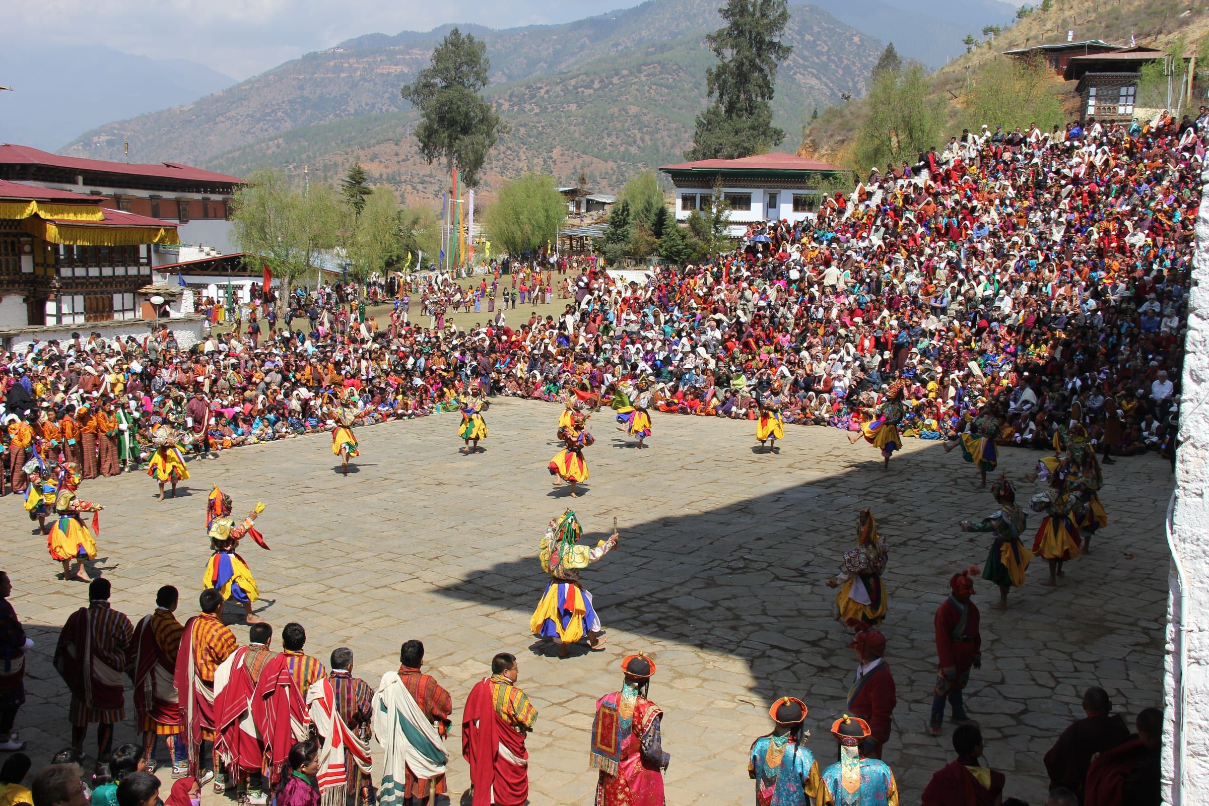 Masked dancers fill a stone courtyard, ringed by a large crowd, with forested hills behind.