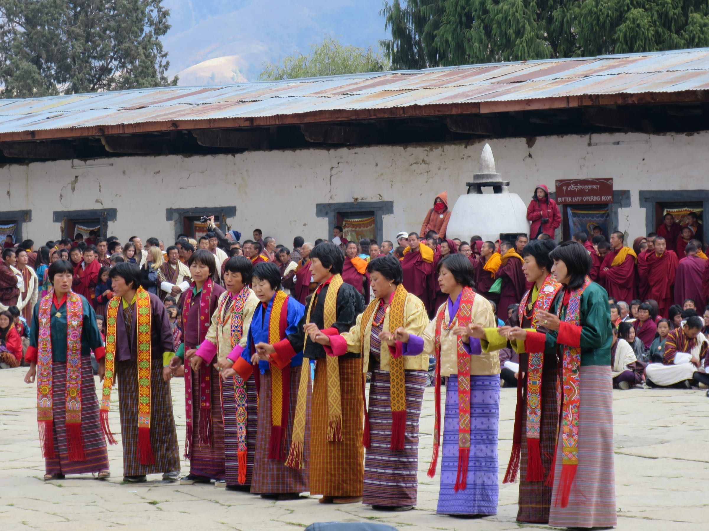 A row of women in kira and rachu perform a folk dance, watched by monks and villagers.