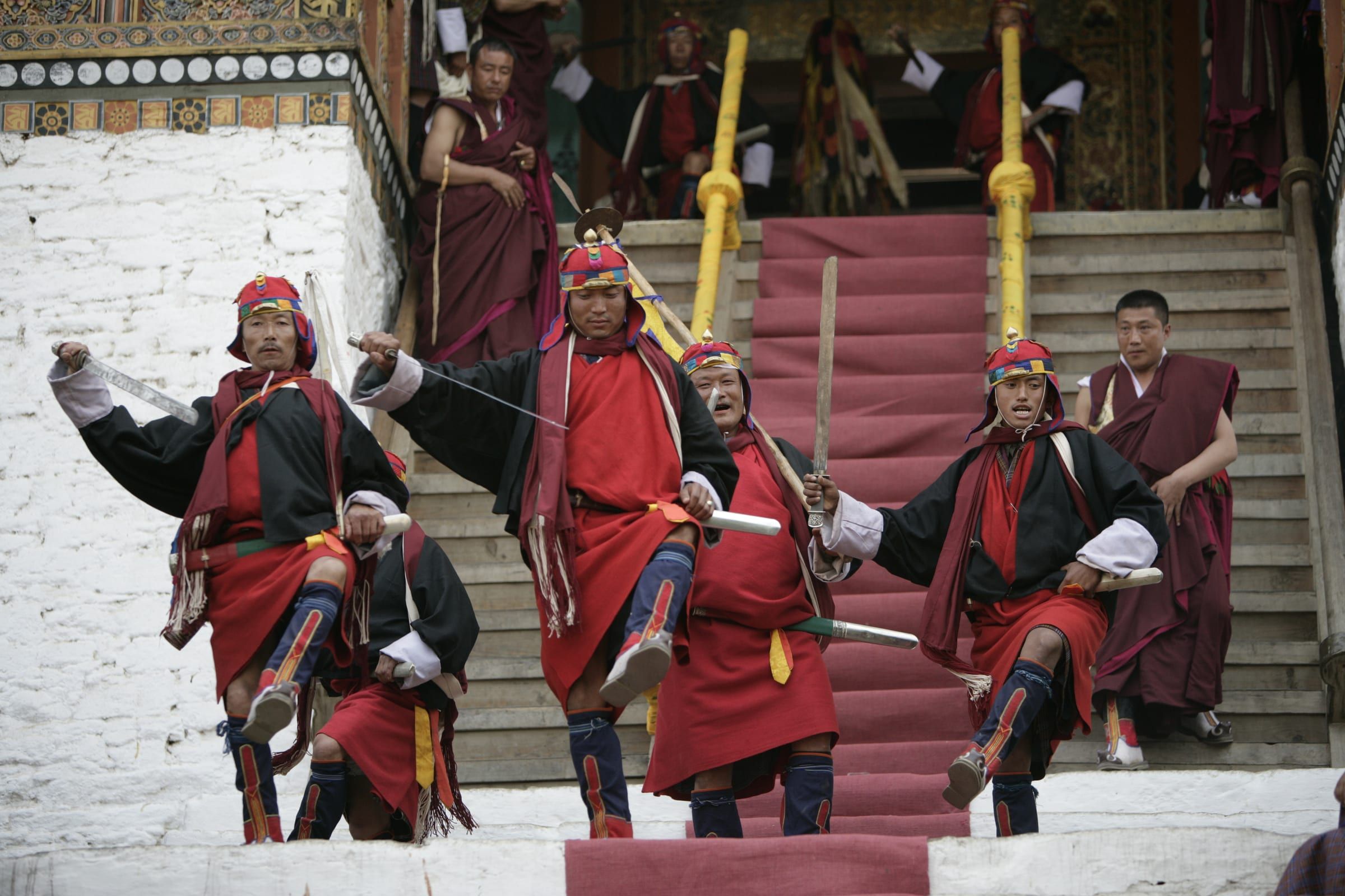 Warriors in red robes dance down the steps of a dzong with raised swords.