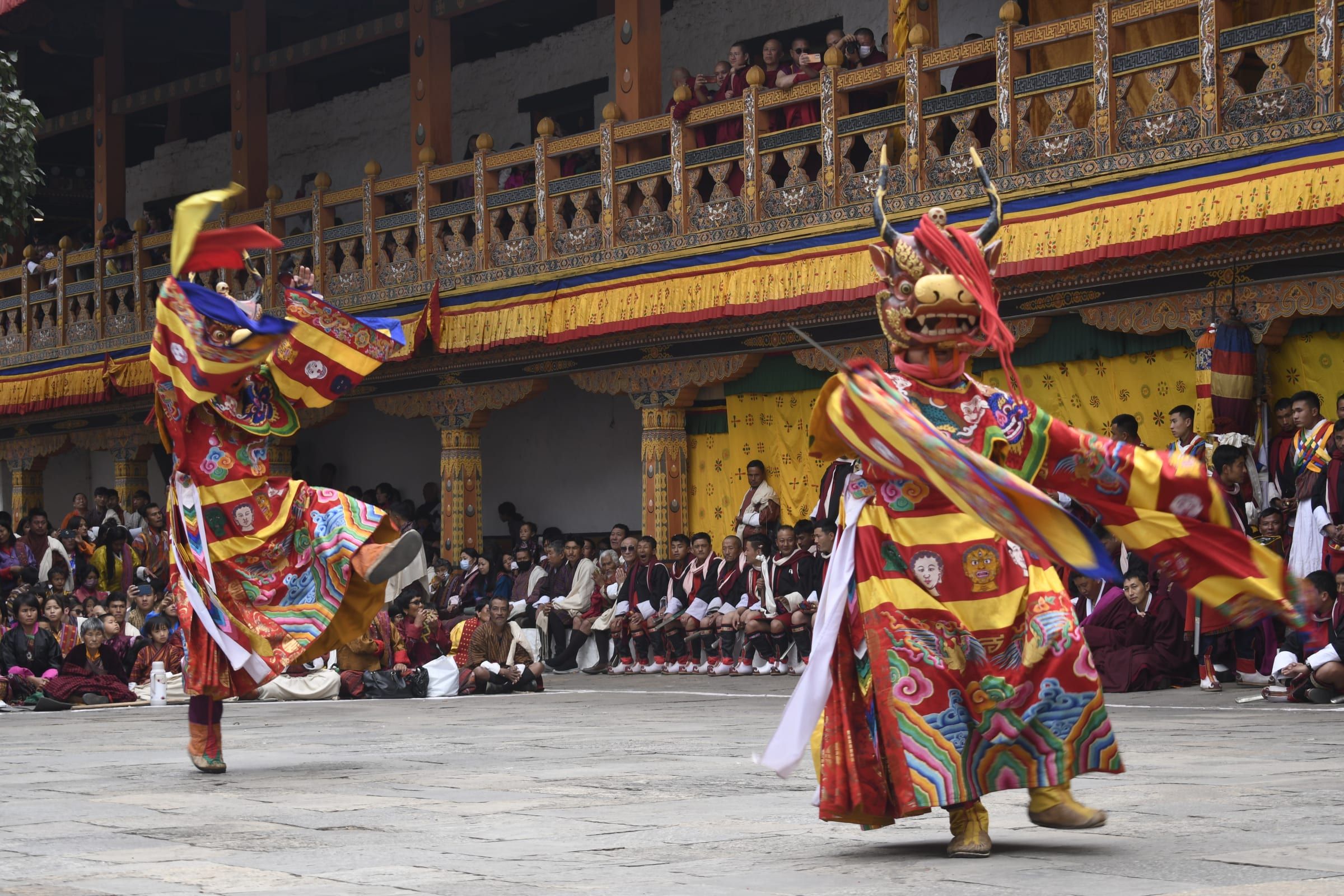 Two masked dancers in brocade whirl in a stone courtyard below crowded wooden balconies.