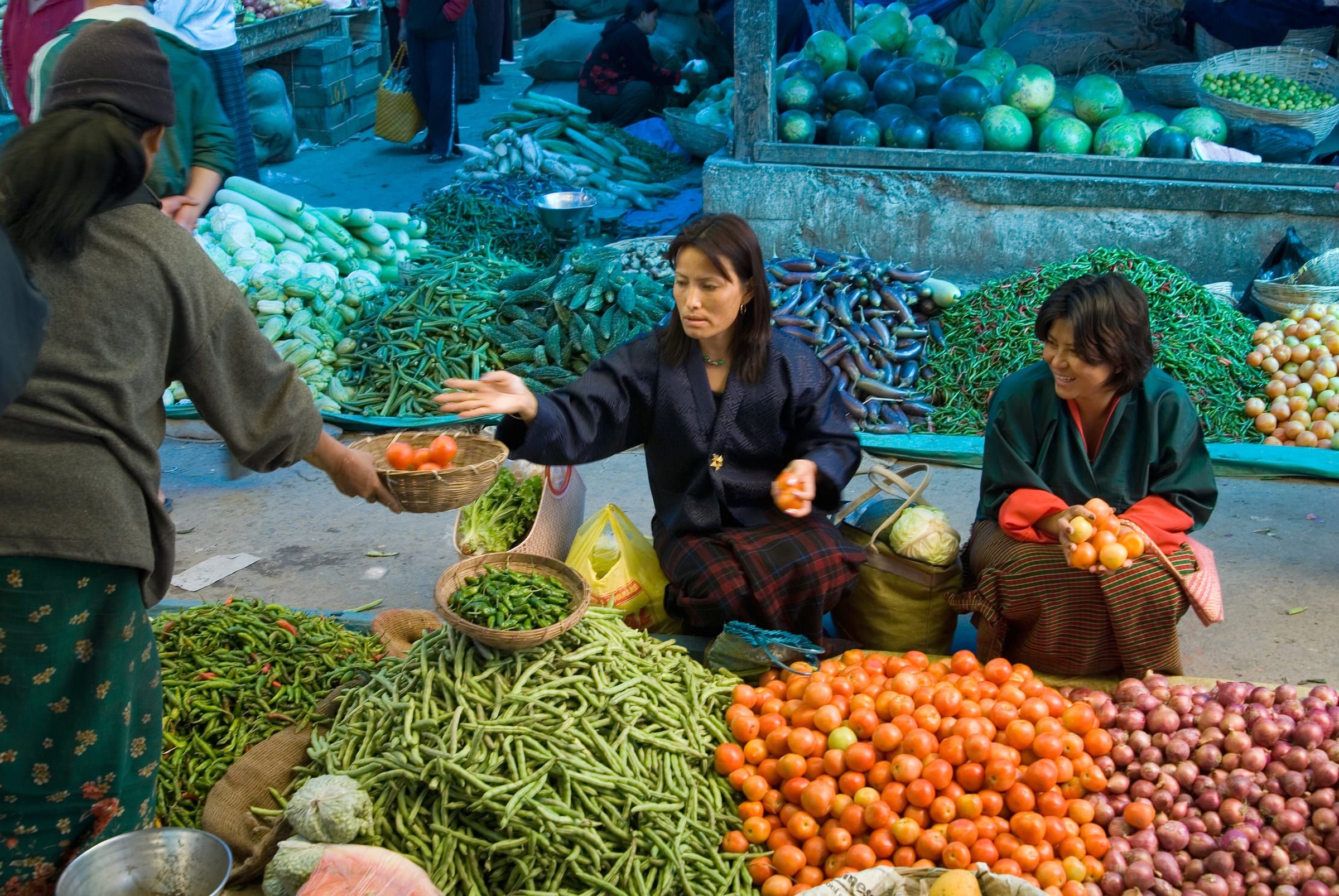 Women in kira sell vegetables from heaped baskets at an outdoor market.