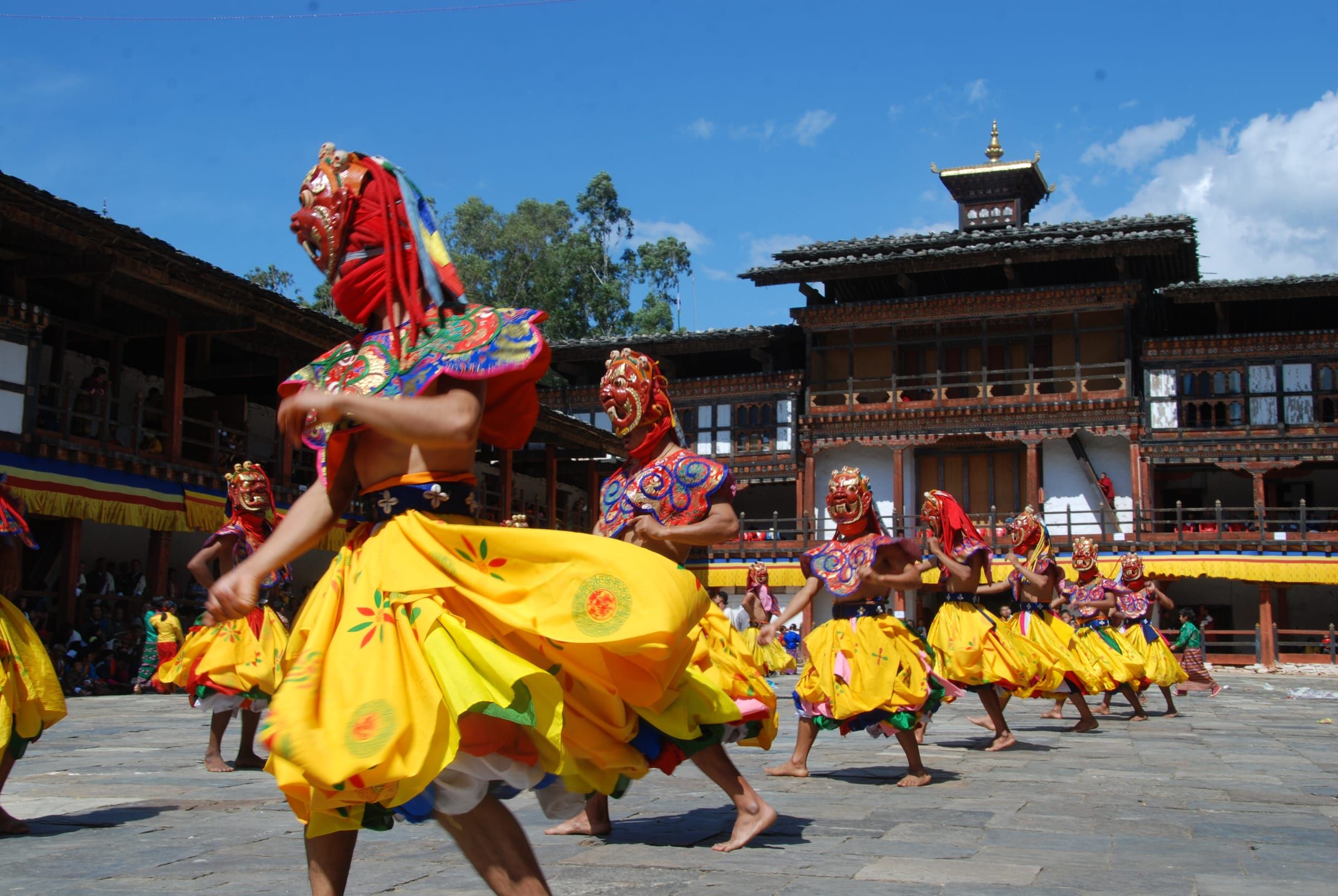 A masked dancer in a flared yellow skirt spins across a flagstone courtyard.