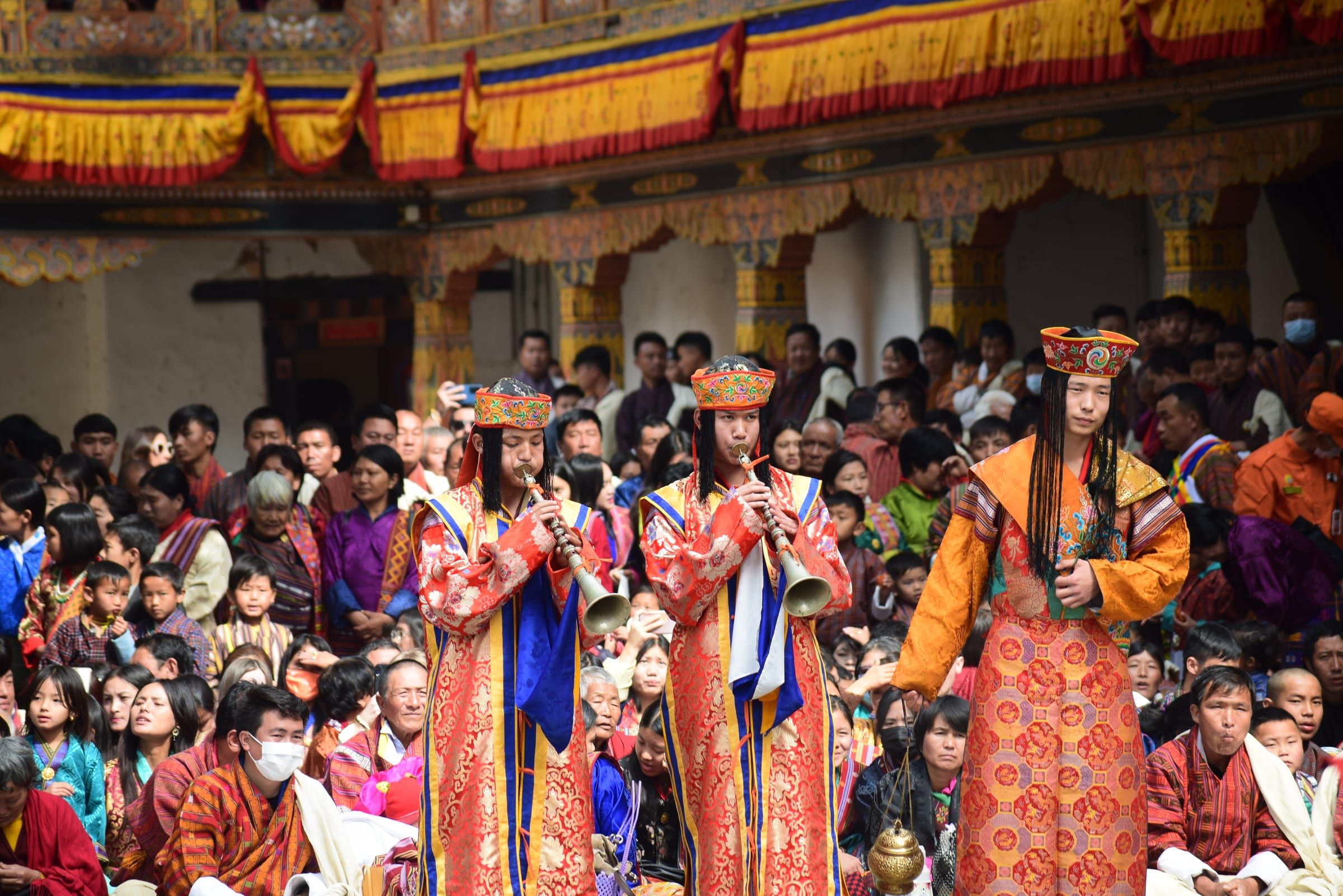 Performers in brocade robes and hats play long horns and carry incense before a crowd.
