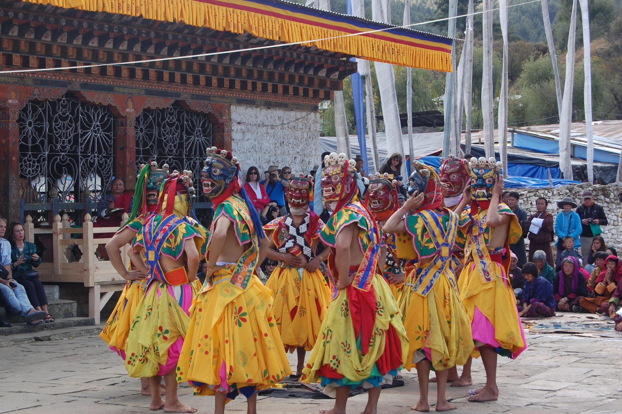 Masked dancers in yellow skirts in a temple courtyard below tall prayer flags.