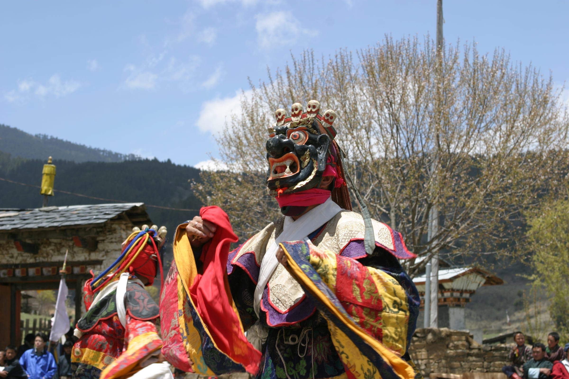 A masked dancer in bright brocade robes and a skull-crowned mask in a village courtyard.