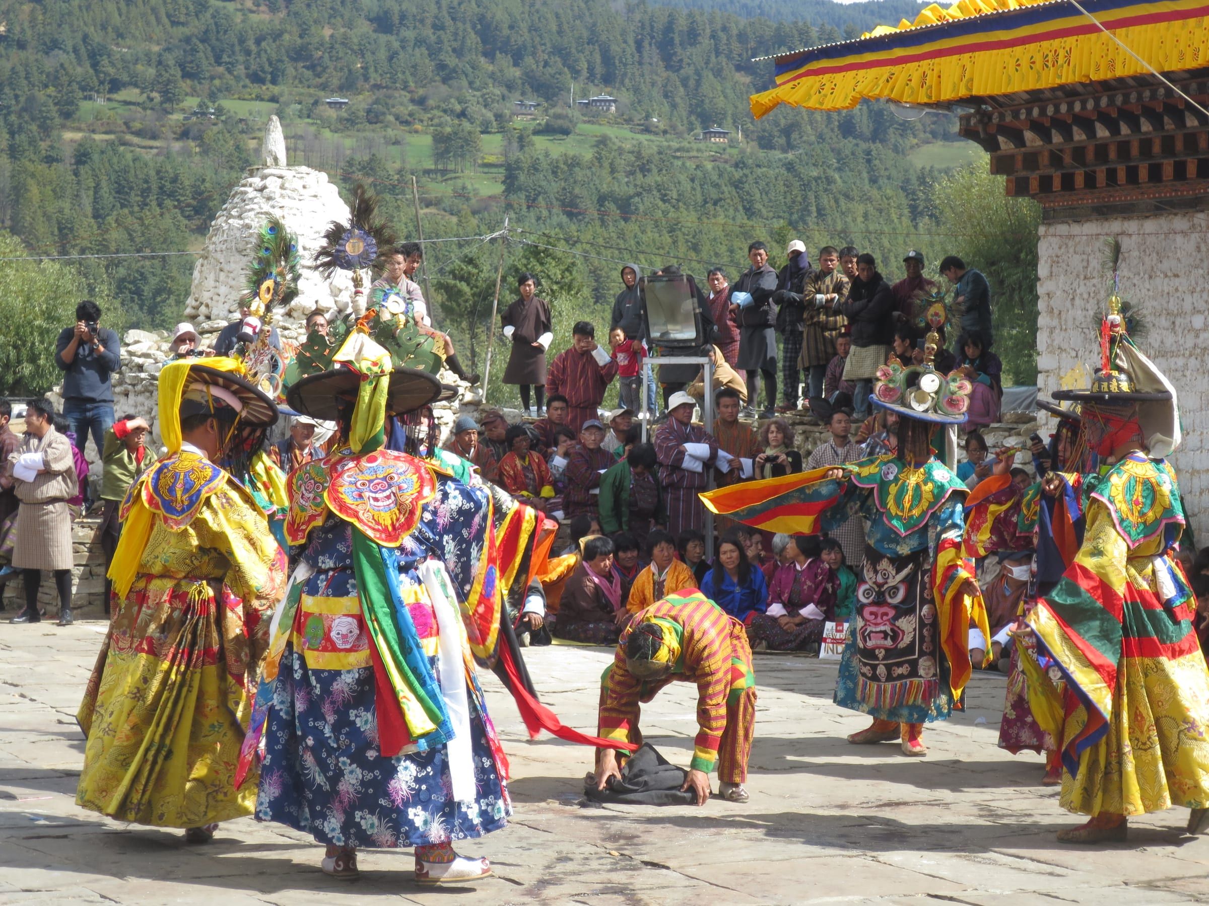Dancers in brocade robes and wide black hats perform before a crowd, with forested hills behind.