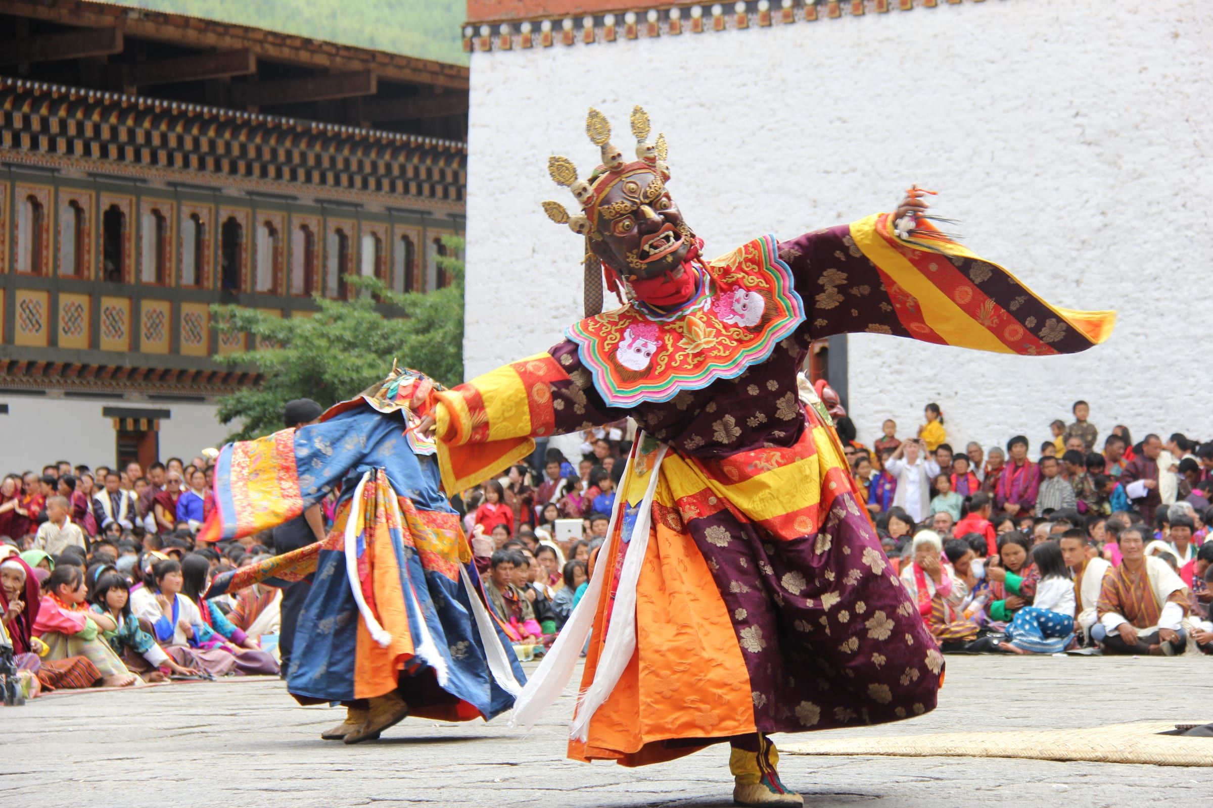 A masked dancer in an ornate robe leaps across a dzong courtyard before a seated crowd.