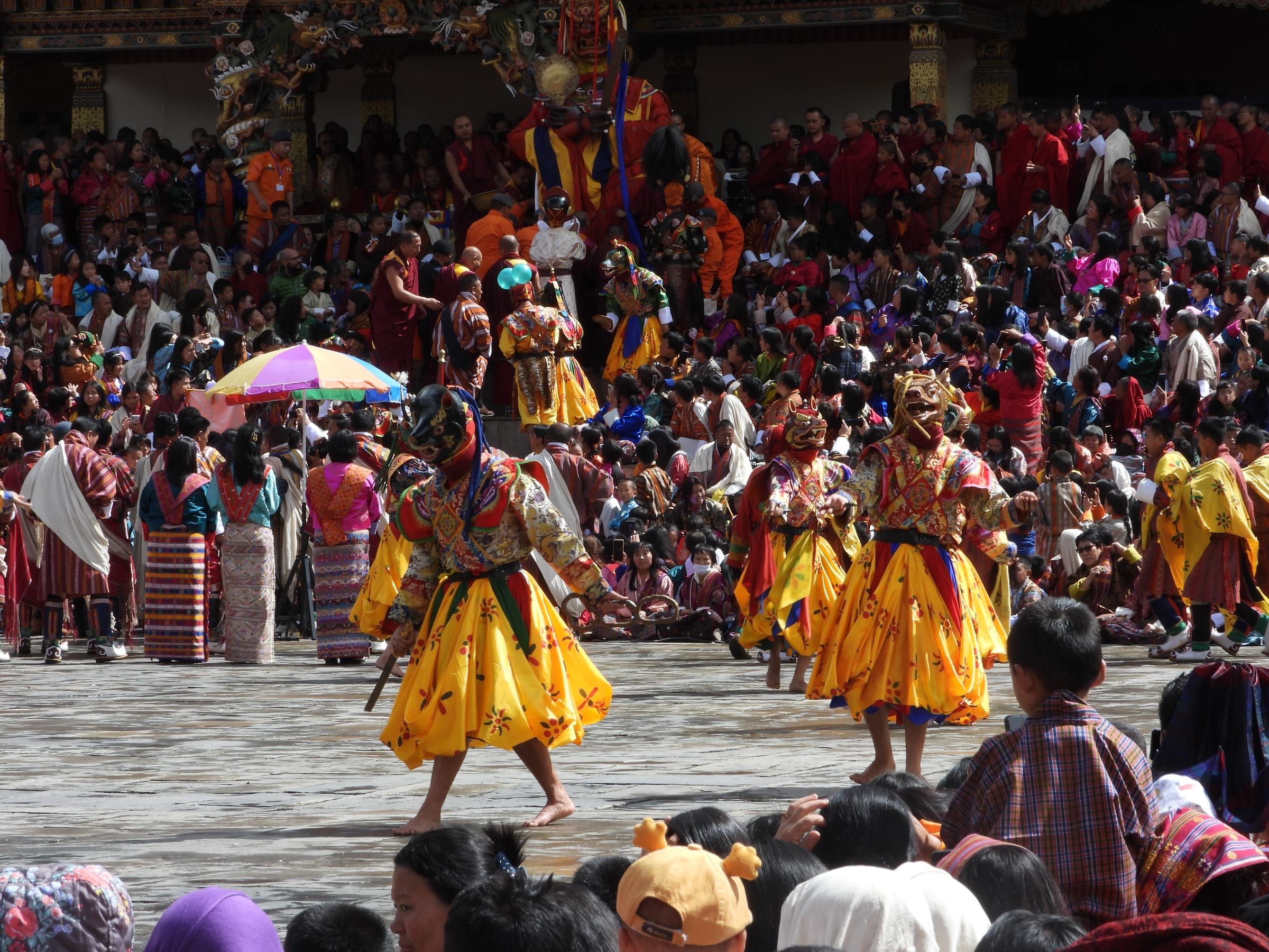 Masked dancers in yellow skirts perform among a dense crowd in a paved courtyard.