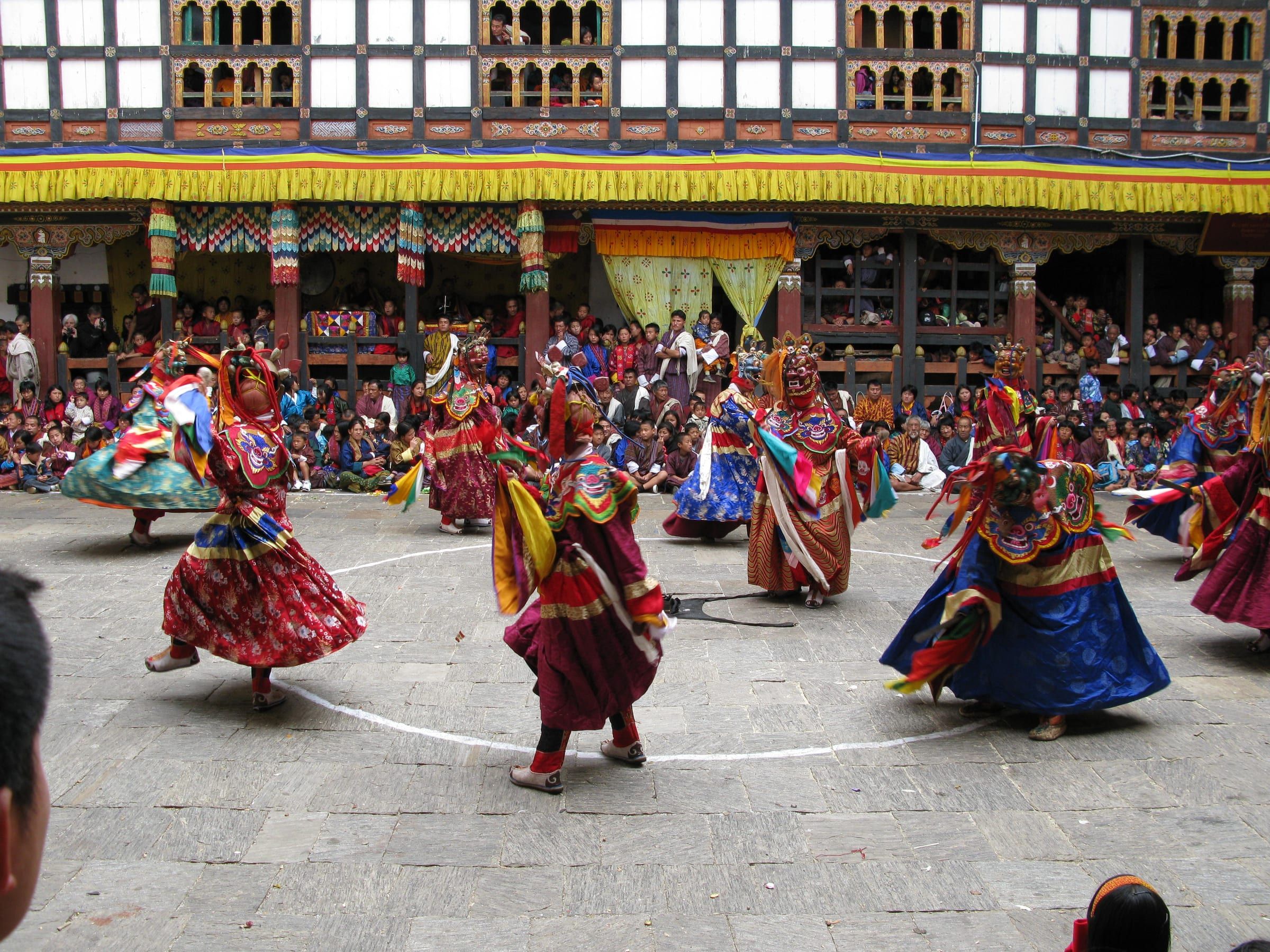Masked dancers in silk robes circle a dzong courtyard, watched from shaded galleries.