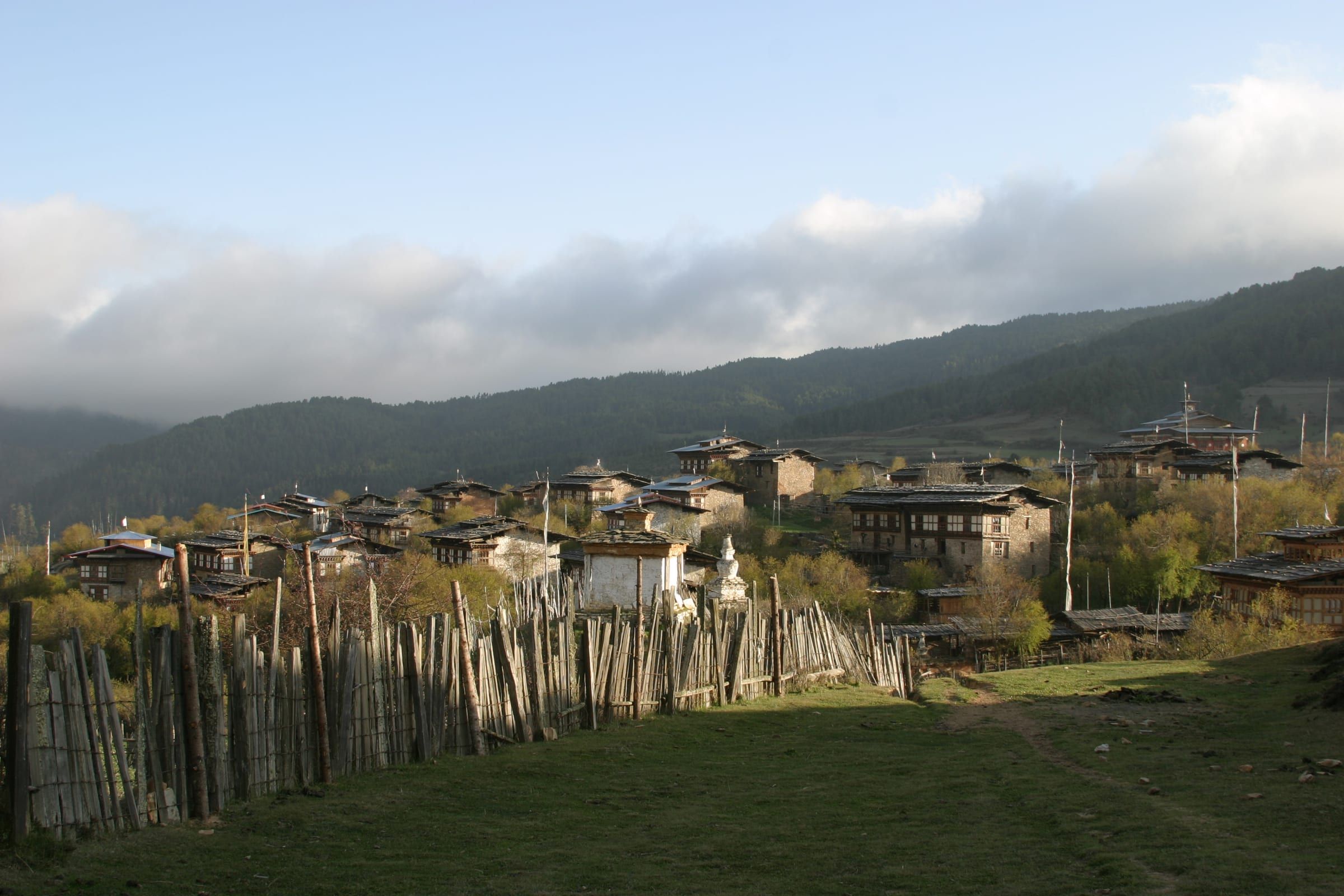 Stone farmhouses behind a wooden fence in a green valley under a cloudy sky.
